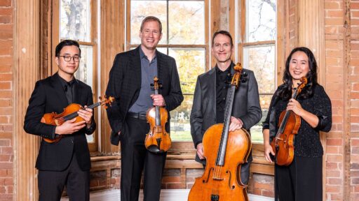 Four members of the New Orford String Quartet stand, holding their instruments, smiling at the camera. Behind them is a window sill with wood panels.
