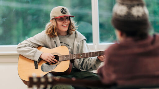 A young adult holds a guitar while facing their instructor. They are wearing a baseball hat and a grey hoodie. They have shoulder length blonde hair.