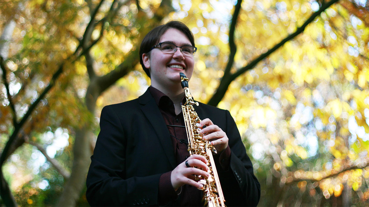 Sylvan Wilkinson smiles while holding a soprano saxophone under a tree canopy