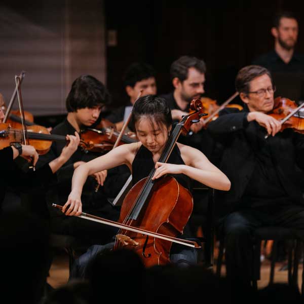 Student soloist plays cello with orchestra behind at Spark to Flame 2025